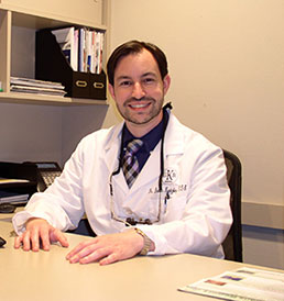 The image shows a man sitting at a desk, smiling towards the camera. He is wearing a white lab coat and appears to be in an office setting.
